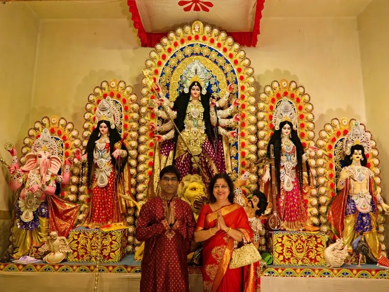 Happy couple tourists in front of Durga Idol pandal - reduced to 40 percent size of Durga Idol centered in front of stage with hands folded namaste - faces kept exactly same no change, dress colours changed to festive maroon red kurta and red saree gold border